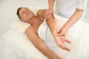 kiropraktik utför en rehabiliterande kropp massage till en stilig mogna caucasian man på wellness spa Centrum. osteopati, fysioterapi, sport skada rehabilitering begrepp, holistiskt vård begrepp foto