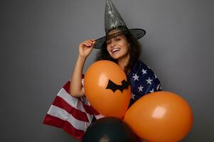 glad blandad lopp ung kvinna i trollkarl hatt, insvept i amerikan flagga, innehar färgrik orange luft bollar, ler toothy leende ser på kamera. halloween begrepp på grå bakgrund med kopia Plats foto