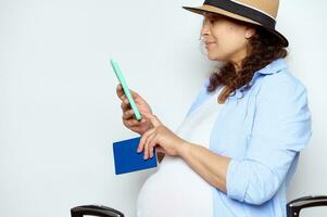 sida porträtt av en gravid kvinna resande i sugrör hatt, kontroll henne mobil telefon, isolerat på vit studio bakgrund foto