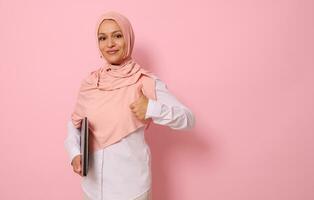 självsäker porträtt av en framgångsrik underbar arab muslim kvinna i rosa hijab och sträng tillfällig klädsel som visar en tumme upp, ser på kamera, Framställ mot färgad pastell bakgrund med Plats för text foto