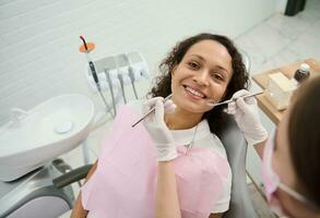 attraktiv afrikansk amerikan kvinna med skön toothy leende och friska vit tänder ler ser på kamera bredvid en tandläkare använder sig av steril dental verktyg under planerad kolla upp i tandvård klinik foto