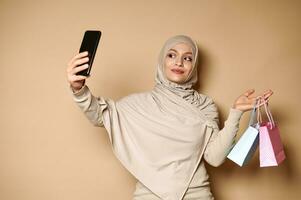 charmig muslim kvinna innehav blå och rosa påsar i ett hand och en smartphone i de Övrig, framställning selfie. handla begrepp foto