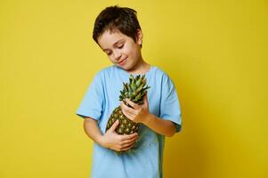 förtjusande barn pojke söt ser på en ananas i hans händer, Framställ över gul bakgrund. sommar och friska vegan mat begrepp foto