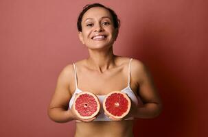 skön kvinna i en vit underkläder innehar grapefrukt halvor på bröst nivå, leende sött med en toothy leende Framställ på en korall bakgrund med Plats för text och reklam. kropp och hud vård foto