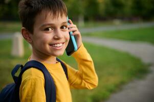 närbild porträtt av en glad vänlig förtjusande skolpojke, i gul tröja talande på mobil telefon, leende med toothy leende ser på kamera på de stad parkera bakgrund foto