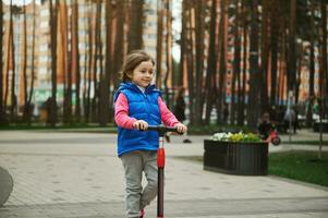 skön europeisk liten flicka, 5 år gammal barn ridning en sparka skoter, njuter solig dag i urban parkera. aktiva och friska livsstil begrepp foto