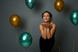 attraktiv sensuell skön kvinna i svart kväll klänning skickar luft kyssar mot grå vägg bakgrund med gyllene och grön metallisk luft ballonger. årsdag, jul, evenemang begrepp, kopia Plats foto