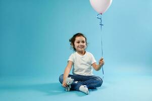 en liten söt flicka sitter på en blå bakgrund och innehar en rosa ballong i ett hand och en teddy Björn mjuk leksak i de Övrig hand foto