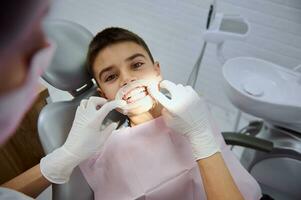 närbild av söt stilig barn skola pojke ser på kamera med skön toothy leende och friska vit tänder medan pediatrisk tandläkare sätta en kind upprullningsdon i hans mun innan dental behandling foto