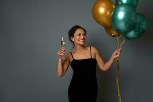 Söt kvinna med skinande gyllene grön metallisk luft ballonger Framställ mot grå vägg bakgrund med kopia Plats innehav en glas av champagne i händer. attraktiv lady firar festlig händelse foto