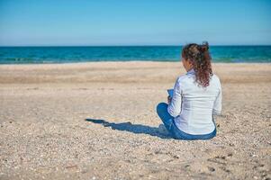 ung brunett sitter på de sand i främre av de skön blå hav och drar på de strand foto