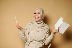 muslim kvinna i hijab innehav en anteckningsblock och penna och skrattande medan Framställ till kamera. beige bakgrund med kopia Plats foto