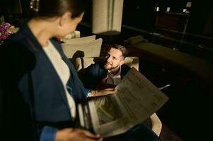 fokus på en stilig caucasian affärsman ser på hans suddig kvinna företag partner, Sammanträde i främre av honom och läsning tidning i en hotell lobby foto