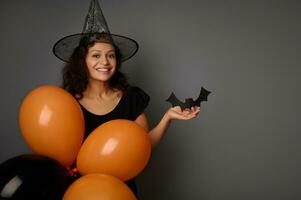attraktiv kvinna i trollkarl hatt, klädd i svart, innehar orange ballonger och handgjort filtskuren svart fladdermus, ler ser på kamera, poser mot grå bakgrund, kopia Plats . halloween fest begrepp foto
