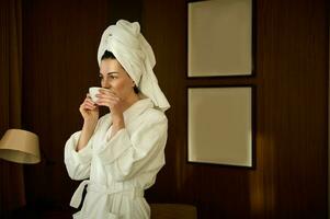 skön självsäker företag kvinna i morgonrock och huvud insvept i handduk njuter de arom av kaffe i de morgon- innan möten under henne företag resa foto