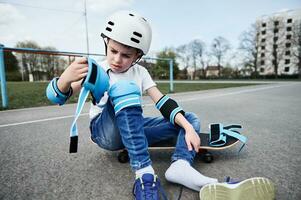 förtjusande pojke skateboarder i säkerhet hjälm och redskap Sammanträde på trä- skateboard på lekplats asfalt foto