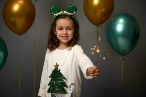 söt bebis flicka med älva ring och Tröja med jul träd innehar sparklers och ler mot grå bakgrund dekoration med lyx gyllene grön skinande luft ballonger. förtjusande barn på ny år fest foto