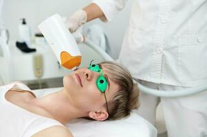 närbild av ung kvinna i skyddande uv laser glasögon tar emot laser behandling på ansikte i samtida spa Centrum foto