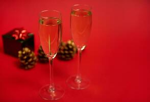 två räfflor av champagne eller glasögon med gnistrande vin på de röd bakgrund med tall koner utsmyckad med gyllene målarfärger och lyx jul gåva i skinande grön omslag papper med glitter röd rosett foto