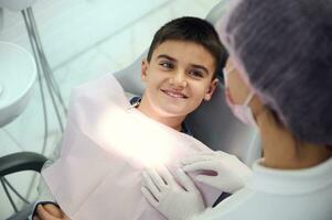 barnläkare tandvård begrepp. dental oral hygien. över huvudet se av caucasian liten pojke leende med skön toothy leende ser på medicinsk assistent framställning honom för regelbunden dental kolla upp foto