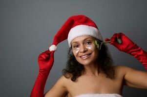 ung skön kvinna klädd som santa användningar jade vält massager för lymfatisk dränering ansiktsbehandling massage, poser på grå bakgrund med kopia Plats för jul annons av skönhet salonger. hud vård foto