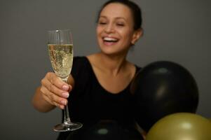 fokus på en gnistrande vin i glas i de hand av en suddig kvinna leende toothy leende innehav guld svart luft ballonger grå bakgrund med kopia Plats för annons. fira svart fredag foto