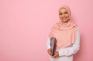 underbar muslim kvinna i eleganta traditionell religiös utrusta med täckt huvud i rosa hijab leende med toothy leende Framställ mot färgad bakgrund med en bärbar dator i händer. kopia Plats foto
