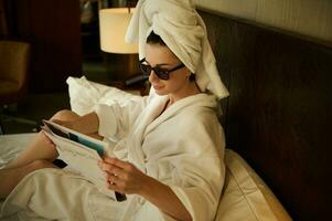över huvudet se av en caucasian Söt kvinna insvept i vit bad handduk läsning Nyheter på tidning och vilar, efter avkopplande badning, på de säng i de hotell sovrum foto