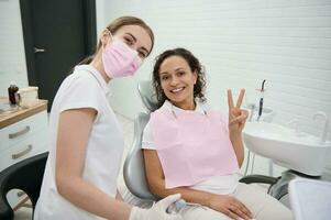 Lycklig afrikansk amerikan kvinna patient som visar en fred tecken Sammanträde i tandläkare stol bredvid en dental praktiker och ler med skön toothy leende ser på kamera efter få dental behandling foto