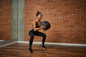 ung afrikansk kvinna med skön muskulös kropp håller på med övningar på de rumpa, hukande med en medicinsk gymnastiska boll medan Träning i de Gym foto