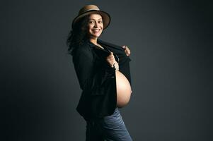 latin amerikan gravid Söt kvinna i sugrör hatt, eleganta blazer, ler på kamera, Framställ bar mage, studio bakgrund foto