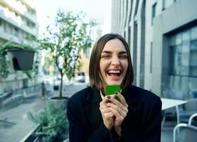 glad förvånad kvinna innehav kreditera kort med grön Chromakey nära henne ansikte, ler toothy leende Framställ mot de bakgrund av modern höghus byggnader bakgrund. företag och finansiera begrepp foto