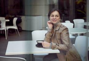 skön attraktiv fantastisk brunett kvinna med en eleganta kort frisyr i en beige dike täcka Sammanträde på en tabell utomhus- och åtnjuter morgon- kaffe med en drömmande se foto
