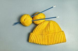 ett Artikel fotografera av en gul stickat ull hatt och två lindning ull trådar strung på lång blå stickning nålar på en grå bakgrund foto