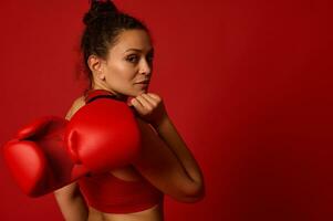 attraktiv vågig håriga Söt kvinna idrottare, sport kvinna boxare kämpe poser mot färgad bakgrund med röd boxning handskar ser självsäkert på kamera genom henne axel. krigisk konst begrepp foto