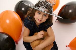 4 år gammal förtjusande Söt liten flicka i häxa klänning och trollkarl hatt utseende på kamera spelar med ballonger och en pumpa isolerat på vit bakgrund. begrepp av en barn har roligt på halloween fest foto