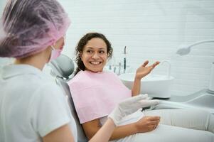 glad afrikansk kvinna Sammanträde i tandläkare stol på planerad regelbunden dental kolla upp i vit tandvård klinik med samtida Utrustning. oral vård och hygien begrepp. dental hygienist samråd foto