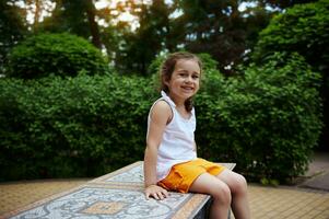 busig liten barn, förskola flicka 4 år leende ser på kamera, Sammanträde på en mosaik- bänk i de stad parkera foto