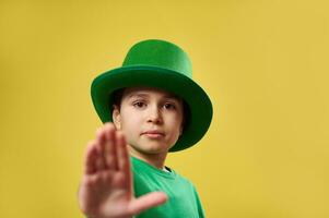 allvarlig liten pojke bär irländsk pyssling grön hatt visar sluta till de kamera. helgon Patricks dag. kopia Plats. gul bakgrund foto