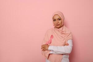 självsäker porträtt av skön mitten östra etnicitet arab kvinna, bär rosa hijab med rosa bröst cancer medvetenhet band, ser på kamera, Framställ över rosa bakgrund med kopia Plats foto