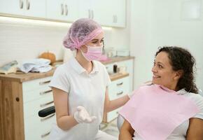 tandläkare hygienist, dental assistent bär rosa medicinsk mask och kirurgisk handskar framställning kvinna patient till medicinsk kolla upp, ger samråd handla om oral hygien i tandvård modern klinik foto