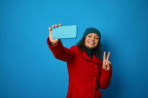 glad skön kvinna bär ljus röd täcka och värma grön ull- hatt innehav mobil telefon i utsträckt hand och som visar fred tecken ser på kamera medan framställning selfie på blå bakgrund foto