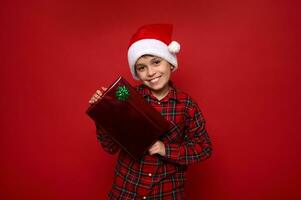 stilig förstadium pojke i santa claus hatt och röd pläd skjorta poser över färgad bakgrund med jul gåva, log med skön toothy leende ser på kamera. kopia Plats för annons foto