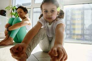 förtjusande liten flicka söt ser på kamera, stretching henne kropp mot de bakgrund av henne mamma under yoga öva foto