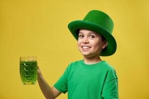leende pojke i grön pyssling irländsk hatt innehar en glas med grön dryck och poser till de kamera. helgon Patricks dag. isolerat på gul bakgrund. foto