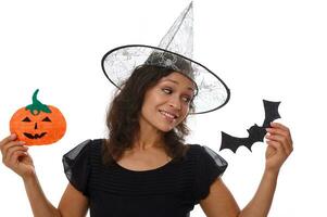 överraskad mystisk leende latinamerikan kvinna i trollkarl hatt, klädd i svart, innehar orange handgjort filtskuren pumpa och fladdermus, poser mot vit bakgrund med kopia Plats för halloween annons foto