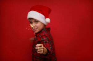 fokus på de brand pärlar i de sparkler bengal lampor i de hand av suddig barn i santa claus hatt och rutig skjorta, isolerat över röd färgad bakgrund med kopia Plats för jul ad foto