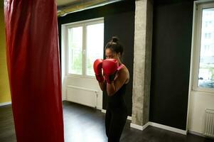 kondition kvinna boxare i TÄTSITTANDE svart sportkläder och röd boxning handskar Träning i en boxning Gym, framställning en hetero stansa slå en stansning väska under ett inomhus- träna foto