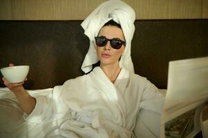 modern eleganta kvinna bär solglasögon och vit frotté morgonrock, omslag huvud i handduk, liggande på säng i sovrum, vilar efter dusch, innehav kopp av varm dryck i ett hand och läsning tidning foto