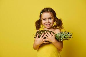 förtjusande liten flicka, klädd i gul, innehav en ananas och leende söt, Framställ i främre av de kamera på en gul bakgrund med kopia Plats foto